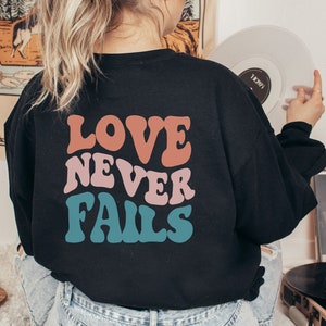 Pode incluir: Uma camisa preta de manga comprida com o texto "LOVE NEVER FAILS" impresso em letras retr&ocirc; coloridas.