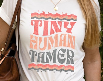 May include: A white t-shirt with a retro-style graphic that says "Tiny Human Tamer" in pink and gray lettering. The graphic is surrounded by a wavy line with stars.