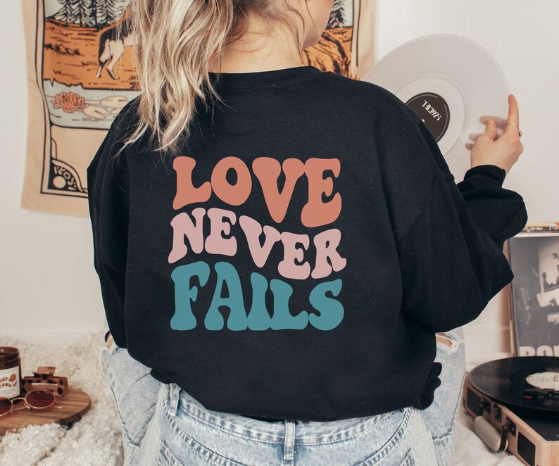 Pode incluir: Uma camisa preta de manga comprida com o texto "LOVE NEVER FAILS" impresso em letras retr&ocirc; coloridas.