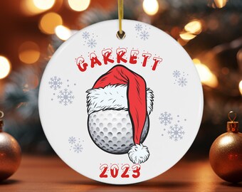 Puede incluir: Adorno de Navidad de cerámica blanca con una bola de golf que lleva un gorro de Papá Noel. El adorno tiene el nombre "GARRETT" y el año "2023" impreso.