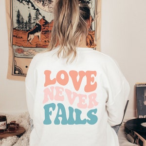 Pode incluir: Camisola branca com o texto "LOVE NEVER FAILS" em letras retro coloridas.