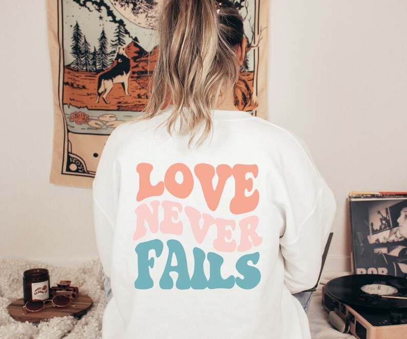 Pode incluir: Camisola branca com o texto "LOVE NEVER FAILS" em letras retro coloridas.