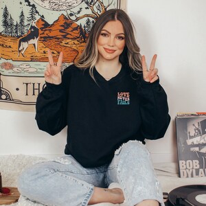 Pode incluir: Uma mulher vestindo uma camiseta preta com o texto "LOVE NEVER FAILS" em cores do arco-&iacute;ris. Ela est&aacute; sentada em uma cama com um toca-discos e uma pilha de discos no fundo.