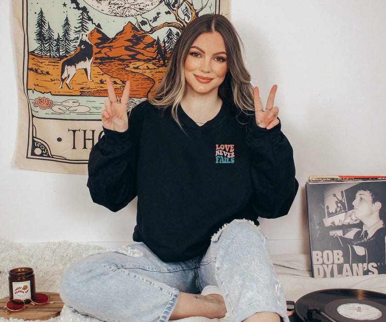 Pode incluir: Uma mulher vestindo uma camiseta preta com o texto "LOVE NEVER FAILS" em cores do arco-&iacute;ris. Ela est&aacute; sentada em uma cama com um toca-discos e uma pilha de discos no fundo.