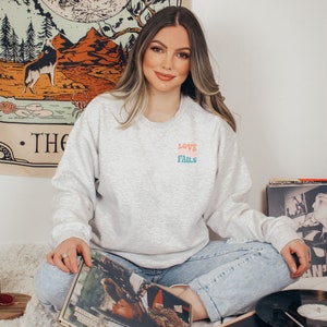 Pode incluir: Uma mulher vestindo uma camiseta cinza com o texto "LOVE FAILS" em letras coloridas. Ela est&aacute; sentada em um tapete branco com dois discos de vinil &agrave; sua frente.