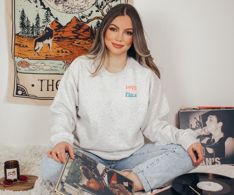Pode incluir: Uma mulher vestindo uma camiseta cinza com o texto "LOVE FAILS" em letras coloridas. Ela est&aacute; sentada em um tapete branco com dois discos de vinil &agrave; sua frente.
