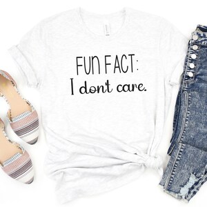 May include: White t-shirt with the text "FUN FACT: I don't care." printed in black. The shirt is laid flat on a white surface with a pair of brown and white striped flats and a pair of blue jeans.