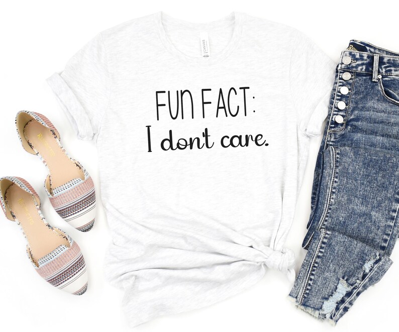 May include: White t-shirt with the text "FUN FACT: I don't care." printed in black. The shirt is laid flat on a white surface with a pair of brown and white striped flats and a pair of blue jeans.