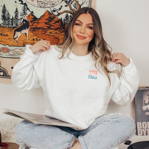 Pode incluir: Uma camiseta branca de manga comprida com as palavras "LOVE NEVER FAILS" impressas em rosa, azul e verde na frente. A camiseta est&aacute; sendo usada por uma pessoa sentada no ch&atilde;o com um toca-discos e um disco na frente.