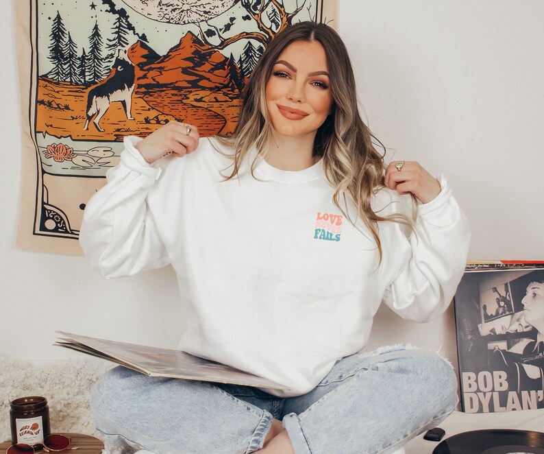 Pode incluir: Uma camiseta branca de manga comprida com as palavras "LOVE NEVER FAILS" impressas em rosa, azul e verde na frente. A camiseta est&aacute; sendo usada por uma pessoa sentada no ch&atilde;o com um toca-discos e um disco na frente.