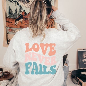 Pode incluir: Uma camisa de moletom cinza com o texto "LOVE NEVER FAILS" em letras coloridas de estilo retr&ocirc;.