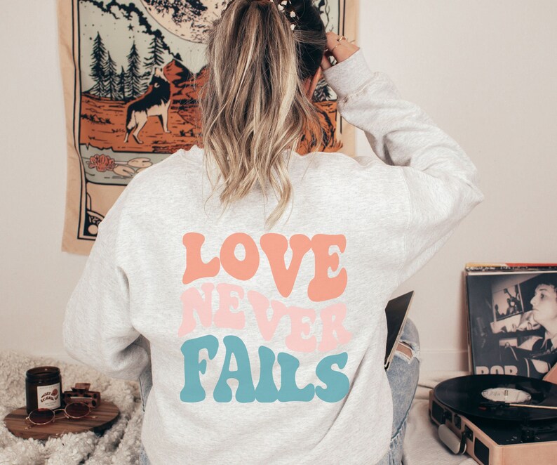 Pode incluir: Uma camisa de moletom cinza com o texto "LOVE NEVER FAILS" em letras coloridas de estilo retr&ocirc;.