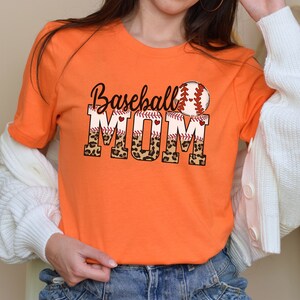 May include: Orange t-shirt with the text "Baseball Mom" in black letters with a red baseball stitch design. The letters "MOM" are partially covered with a leopard print pattern. A red baseball with white stitching is above the text.