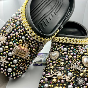 Könnte beinhalten: Schwarze Clogs, verziert mit goldenen Ketten, Perlen, Strasssteinen und Charms. Die Charms beinhalten ein Parfümfläschchen mit dem Text "You are beautiful", ein Herz, eine Krone und einen Liebescharm.