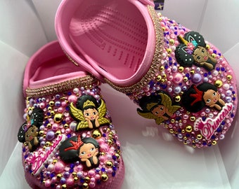 Puede incluir: Crocs rosas decoradas con diamantes de imitación, perlas y dijes con imágenes de dibujos animados de niñas negras con alas y coronas. Las Crocs tienen la palabra "Princess" escrita en ellas.