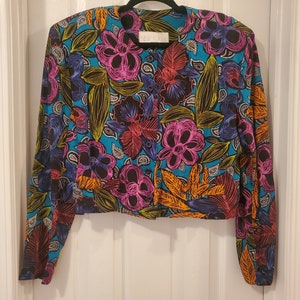 May include: A cropped, long-sleeved jacket with a vibrant floral print. The jacket features a blue background with pink, orange, yellow, and green flowers. The jacket has a button closure and a collar.