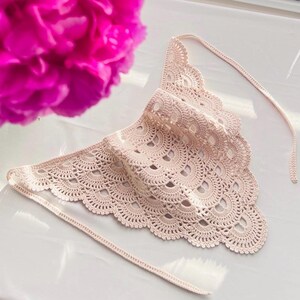 May include: A light beige triangular crochet bandana featuring a delicate scalloped edge pattern with openwork details. The accessory includes two long, thin crocheted ties extending from the top corners, crafted with a consistent stitch design.