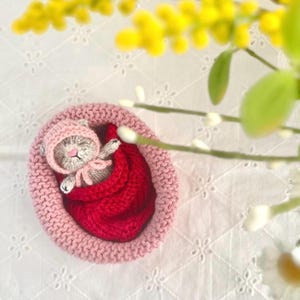 May include: A small, gray, crocheted animal figure wearing a pink bonnet, tucked into a bright red crocheted blanket. This figure rests inside a round, light pink, knitted basket with a textured edge, creating a soft and compact display.