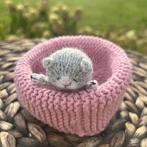 May include: A small, hand-knitted grey kitten figurine with a white muzzle and pink nose, resting inside a round, soft pink knitted basket. The kitten has visible stitched paws and ears, and the basket features a textured, ribbed knit pattern.