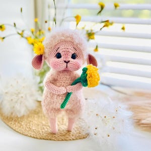 May include: A light pink crocheted lamb figurine with a fuzzy texture, dark round eyes, and a small stitched nose. The lamb has a yellow flower tucked behind one ear and holds a green-stemmed yellow flower in its small crocheted hands.