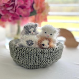 May include: A round, sage green crocheted basket containing four small, fuzzy, handmade kitten figurines. The kittens feature soft, textured yarn in shades of white, gray, and light brown, each with tiny pink noses and dark eyes, nestled together inside the basket.