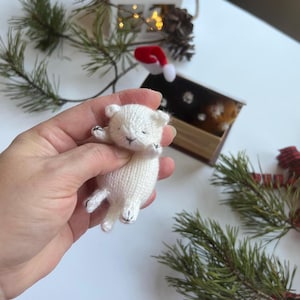 May include: A small, white, hand-knitted toy cat with embroidered black facial features and paw details. The miniature feline figure is crafted from soft yarn, featuring a rounded head, pointed ears, and a slender body, held in a palm.