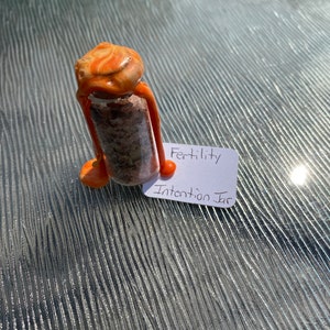 May include: A small glass jar with a brown and orange wax seal. The jar is filled with dried herbs and flowers. A white label on the jar reads "Fertility Intention Jar".
