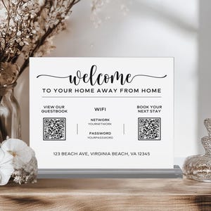 May include: A rectangular white acrylic sign with black text and two QR codes. The sign reads Welcome to your home away from home, View our guestbook, Wifi, Network, Password, Book your next stay, and 123 Beach Ave, Virginia Beach, VA 12345.