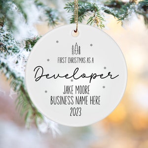 May include: A white ceramic Christmas ornament with the text "FIRST CHRISTMAS AS A Developer" in black script. Additional text includes a name, business name, and the year 2023. The ornament is hanging from a gold string, with a blurred background of evergreen branches.