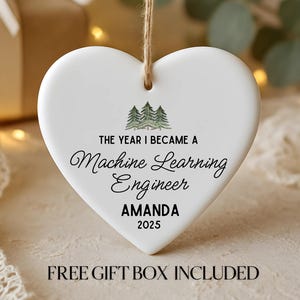May include: White heart-shaped ornament with a string for hanging. The ornament features text that reads "THE YEAR I BECAME A Machine Learning Engineer AMANDA 2025". A small illustration of pine trees is above the text.