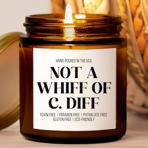 May include: A brown glass candle jar with a white label that reads "NOT A WHIFF OF C. DIFF". The label also states "HAND-POURED IN THE USA", "TOXIN FREE | PARABEN FREE | PHTHALATE FREE", "GLUTEN FREE | ECO-FRIENDLY".