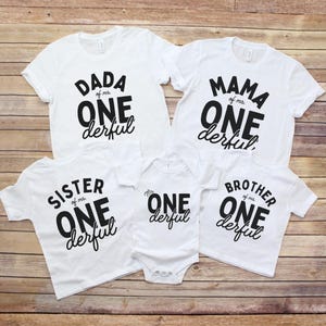 May include: White t-shirts and a onesie with black text that reads "DADA of mr ONE derful", "MAMA of mr ONE derful", "SISTER of mr ONE derful", "mr ONE derful", and "BROTHER of mr ONE derful". The shirts are arranged on a wooden surface.