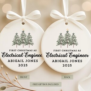 May include: Two white ceramic Christmas ornaments with a ribbon. Each ornament features a watercolor illustration of three green pine trees and the text "FIRST CHRISTMAS AS Electrical Engineer ABIGAIL JONES 2025".
