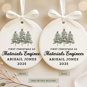 May include: Two white ceramic Christmas ornaments with a watercolor illustration of three evergreen trees. The text reads "First Christmas as Materials Engineer Abigail Jones 2025". Each ornament is tied with a white ribbon.