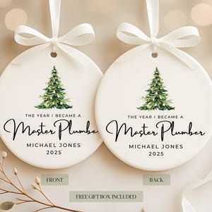 May include: Two white ceramic Christmas ornaments with a green watercolor Christmas tree design. The text reads "THE YEAR I BECAME A Master Plumber" with the name "MICHAEL JONES" and the year "2025". Each ornament has a white ribbon.