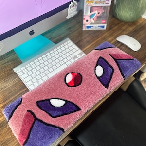 May include: A rectangular, pink rug featuring a cartoon character design with purple, red, and white accents. The rug is placed on a wooden surface near a computer keyboard and mouse. The design includes large eyes and a circular red detail.