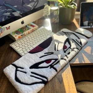 May include: A rectangular, white, gray, and black rug featuring a stylized anime character's face with red eyes. The rug is placed on a wooden desk near a keyboard and computer monitor.