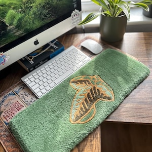 Peut inclure: Un tapis de souris vert avec un motif de feuille brodé en or. Le tapis de souris est sur un bureau en bois avec un clavier, un écran d'ordinateur et une plante en pot.
