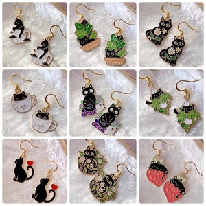 Peut inclure: Une collection de boucles d'oreilles pendantes sur le thème des chats, de couleur dorée. Les motifs incluent des chats noirs dans des tasses, des plantes en pot et des compositions florales. Certaines boucles d'oreilles présentent des chats avec des marguerites, des livres ou des fraises.
