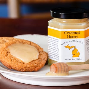 May include: A jar of creamed honey with a black lid and a label that reads "Creamed Honey". Two cookies with a honey glaze sit on a white plate, along with a wooden honey dipper. The jar is labeled "Grand Rapids Bee Co."