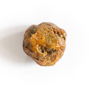 May include: A natural, irregularly shaped stone with a rough, textured surface. The stone displays a mix of brown, tan, and golden-yellow hues, with areas of darker coloration. The object is set against a plain white background.