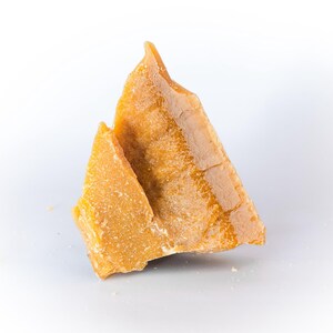 May include: Two pieces of golden-yellow beeswax, with a rough, textured surface. The wax is in irregular, triangular shapes, and appears to be freshly cut. The background is plain white.