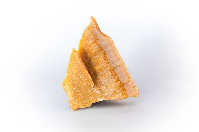 May include: Two pieces of golden-yellow beeswax, with a rough, textured surface. The wax is in irregular, triangular shapes, and appears to be freshly cut. The background is plain white.