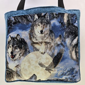 May include: A denim tote bag featuring a fabric panel with a wolf and eagle design. The panel depicts wolves in a snowy landscape with a full moon and an eagle in flight. The bag has black handles.