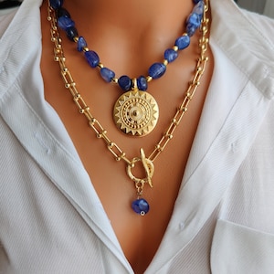 May include: A layered gold chain necklace with a large gold pendant featuring a sunburst design. The necklace is paired with a blue stone beaded necklace.