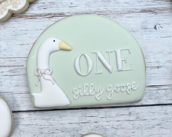 Può includere: Un biscotto di zucchero a forma di semicerchio con base verde pallido, caratterizzato da un'illustrazione di un'oca bianca, un piccolo fiocco rosa e il testo "ONE silly goose" in glassa bianca. Il biscotto è decorato con glassa liscia.