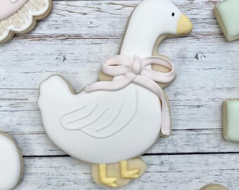 Può includere: Un biscotto di zucchero a forma di oca bianca decorato con un fiocco in pasta di zucchero rosa chiaro intorno al collo, becco e zampe gialle e dettagli delle ali in rilievo. Il biscotto è circondato da altri, tra cui un numero uno verde chiaro.