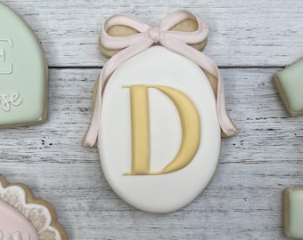 Può includere: Un biscotto ovale decorato con glassa bianca, una lettera D gialla al centro e un fiocco di glassa rosa chiaro sulla parte superiore. Il biscotto è circondato da altri biscotti decorati nei toni del verde salvia, bianco e rosa su legno.