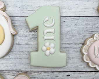 Può includere: Un biscotto color verde salvia chiaro a forma di numero uno, con scritte bianche in rilievo che compongono la parola one, decorato con un piccolo fiore bianco con centro giallo alla base del numero, circondato da altri biscotti.