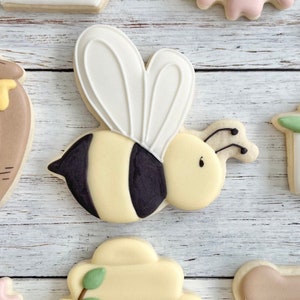 May include: A bee-shaped sugar cookie decorated with royal icing. The cookie features a pale yellow body with two distinct black stripes, large white wings with piped outlines, a small black dot for an eye, and two thin black antennae.
